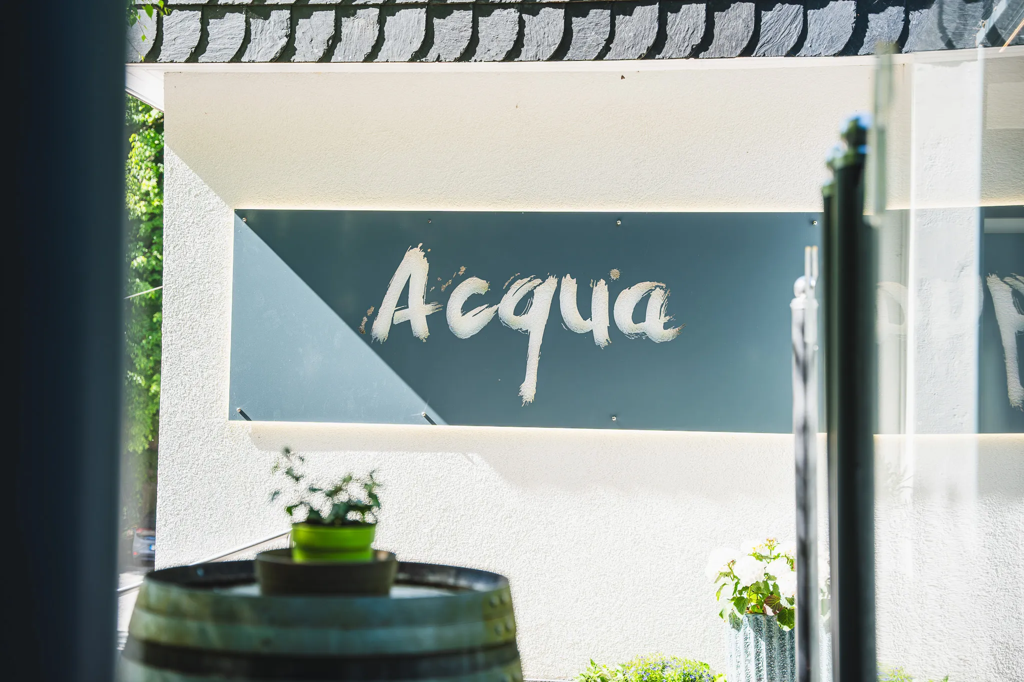 Acqua-Schild im Eingangsbereich, daneben ein Holzfass mit Bepflanzung.