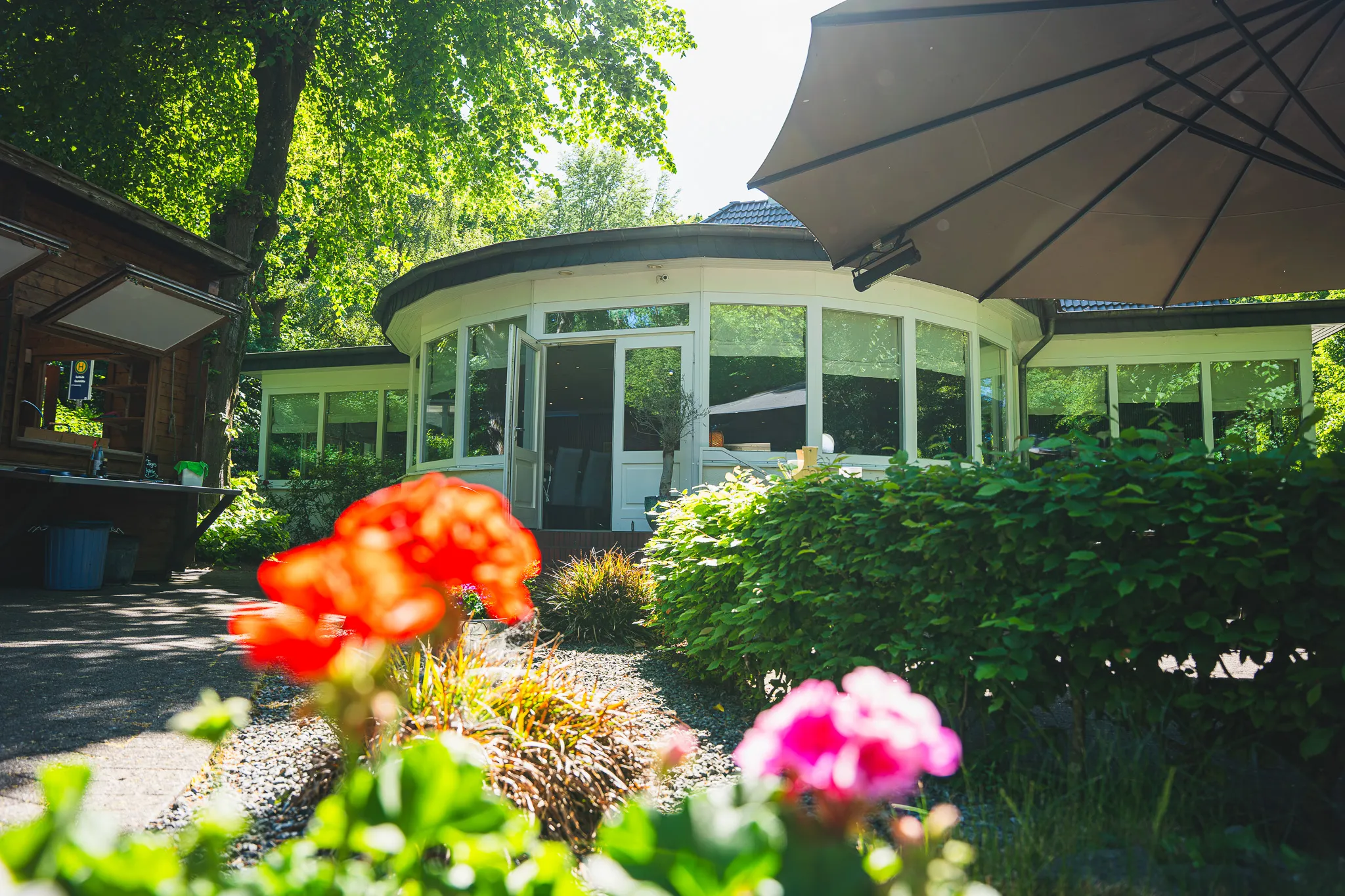 Außenansicht des Restaurants mit Sonnenschirmen, im Vordergrund rote und pinke Geranien.