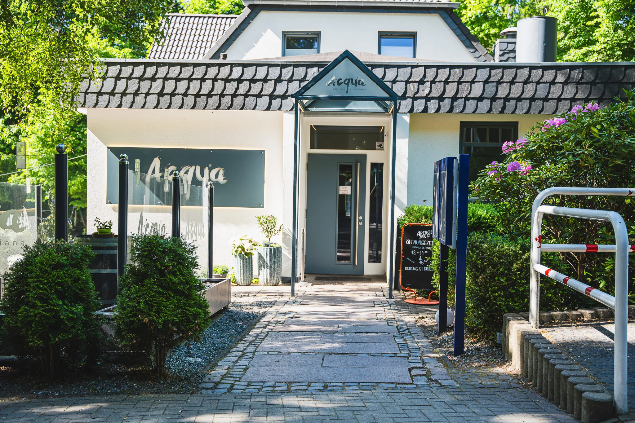 Eingang des Ristorante Acqua: weißes Gebäude mit Schindeldach, Acqua-Schriftzug über der Tür und gepflastertem Weg.