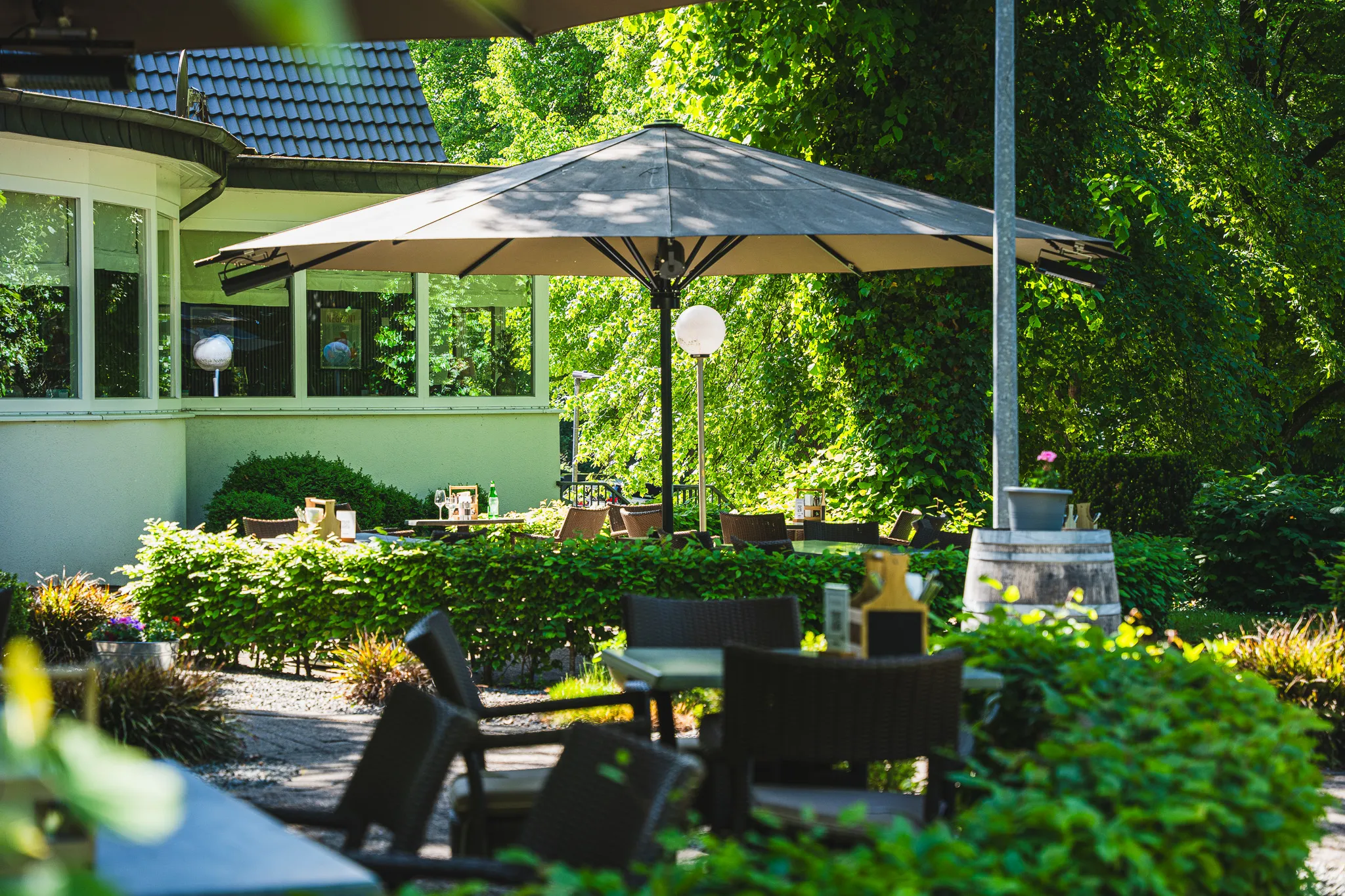 Terrasse im Grünen mit Sonnenschirm, Sitzgruppen und dichtem Baumbestand.